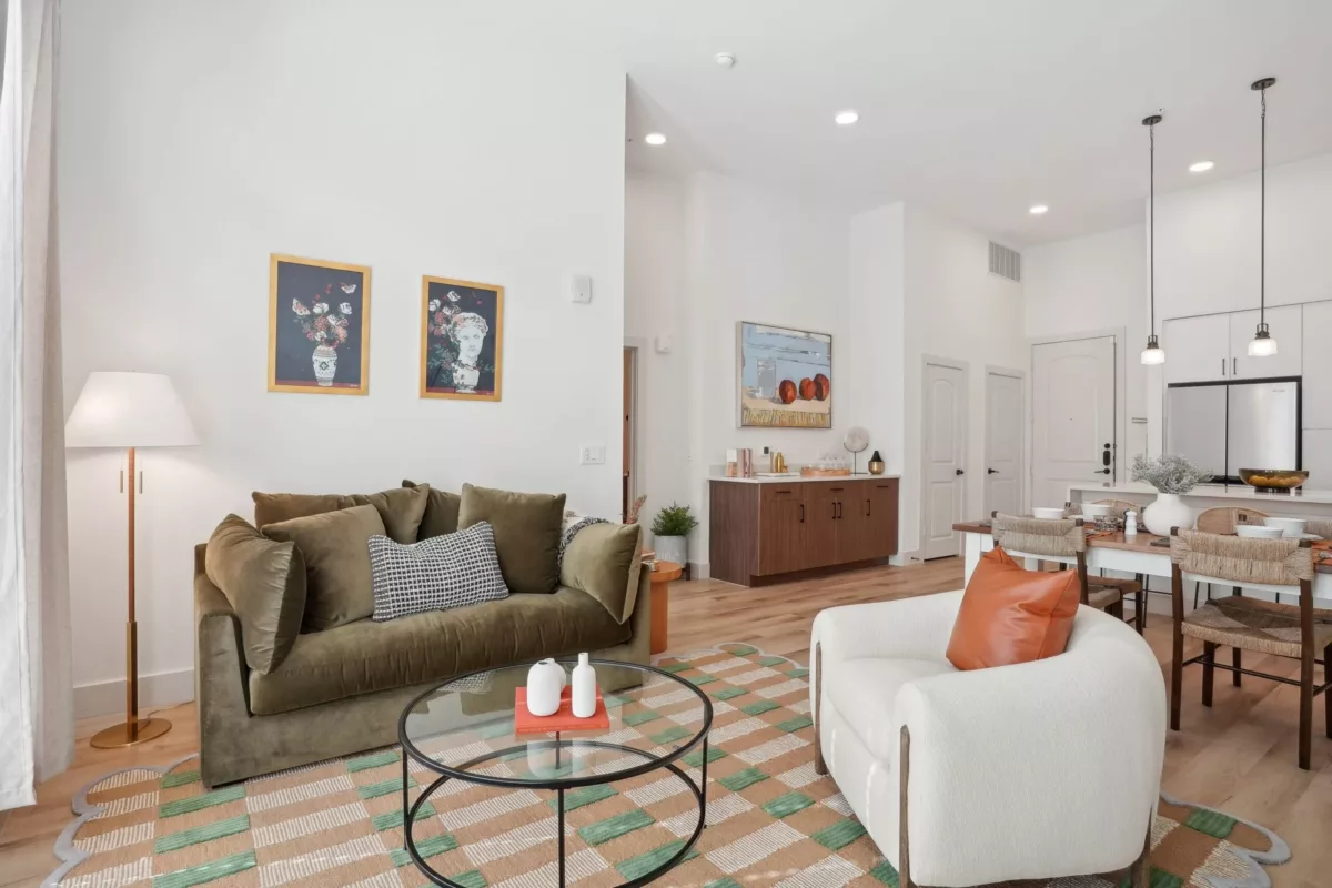 Apartment living room with a green couch, white chair, wood-style flooring with an orange and green-striped rug, and white walls with modern art.