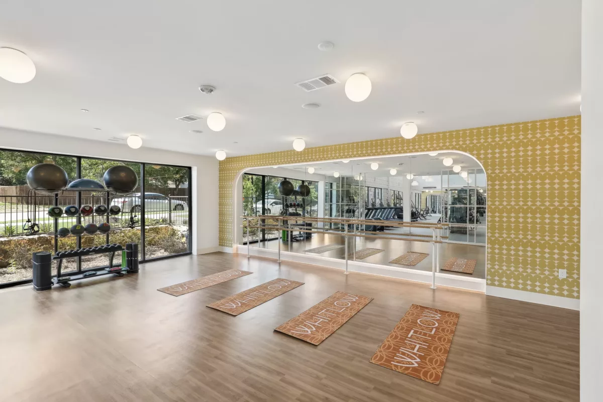 Well-equipped fitness room with yoga mats and a mirrored wall, next to a bright window.