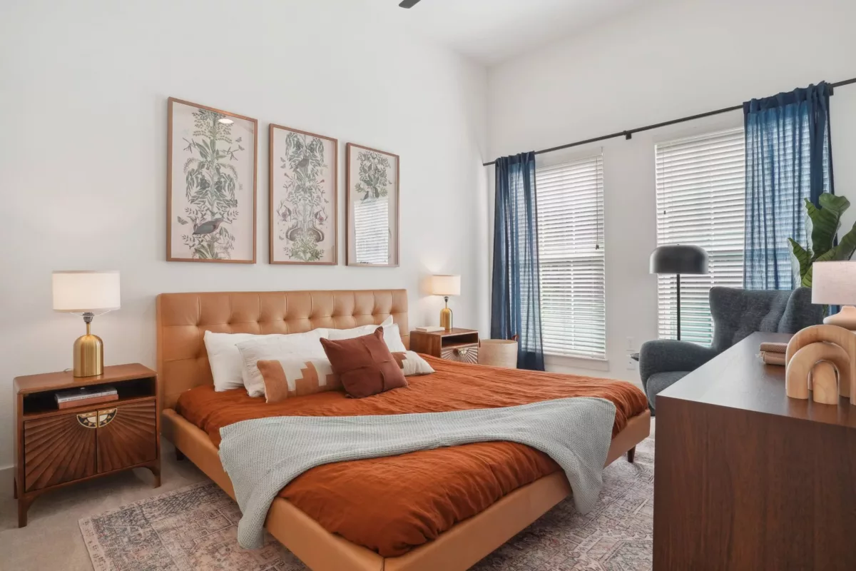 Bedroom with bed with upholstered headboard and orange bedspread, large windows with blue curtains, and artwork on the walls.