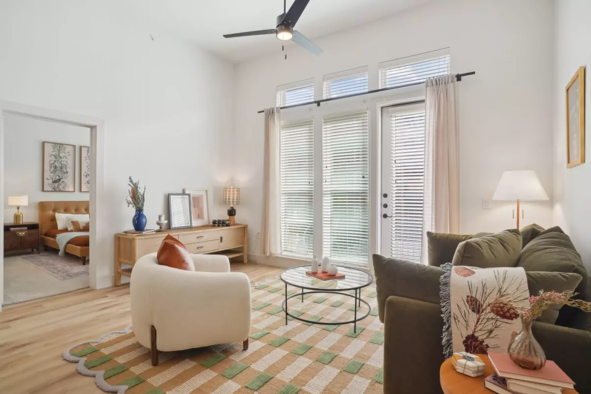 Apartment living room with white walls, ceiling fan, white arm chair, coffee tables, and large windows.
