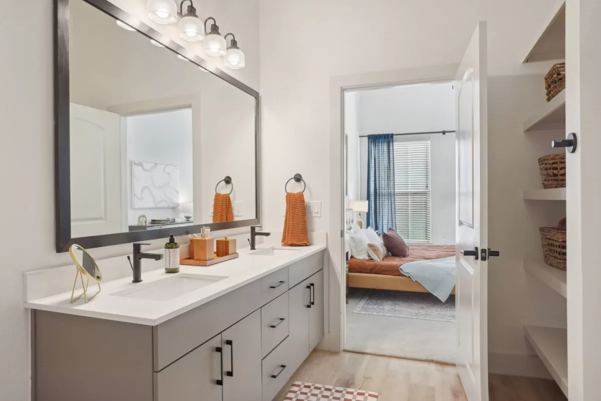 Modern bathroom with dual-sink vanity, large mirror, and view into bedroom with bed and window.